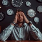 A stressed man holding his head in frustration while surrounded by numerous clocks, symbolizing the challenge of managing time effectively. This image reflects the need for effective time management tips and techniques.